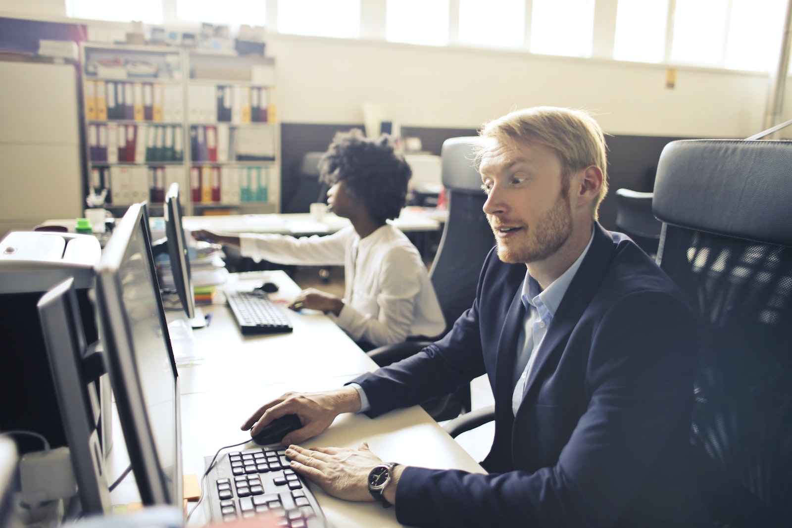 High angle of pensive shocked businessman in elegant clothes focusing on screen and interacting with computer while sitting at table near African American female coworker in light spacious office