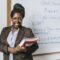 Cheerful black female teacher with workbooks standing near whiteboard